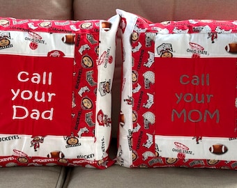 May include: Two red and white decorative throw pillows with the text "Call your Dad" and "Call your Mom" embroidered on them. The pillows feature a patchwork design with the Ohio State logo and the word "Buckeyes" repeated throughout.