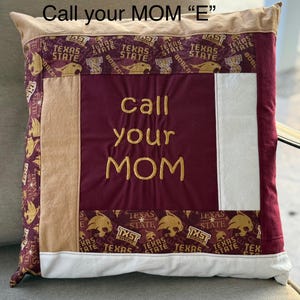 May include: A maroon and tan pillow with a white panel in the center. The pillow has a gold embroidered message that says "Call your MOM" on the maroon panel. The tan panels have a repeated pattern of a maroon and gold Texas State Bobcat mascot.
