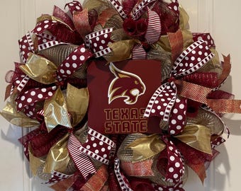 May include: A decorative wreath featuring maroon and gold mesh, accented with ribbons in maroon and white polka dots and stripes. A central square sign displays the Texas State University bobcat logo and the text Texas State in gold lettering.