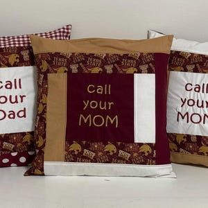 May include: Five decorative throw pillows with a patchwork design. The pillows are made of a variety of fabrics, including red, white, and brown. The pillows have embroidered text that reads "Call your Dad" and "Call your Mom".