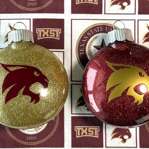 May include: Two round glass ornaments with silver caps. One ornament is covered in gold glitter with a maroon bobcat logo, and the other is covered in maroon glitter with a gold bobcat logo. Both feature a smooth, reflective surface finish.