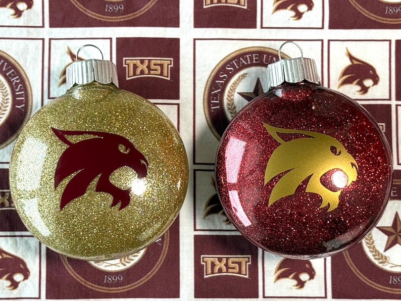 May include: Two round glass ornaments with silver caps. One ornament is covered in gold glitter with a maroon bobcat logo, and the other is covered in maroon glitter with a gold bobcat logo. Both feature a smooth, reflective surface finish.
