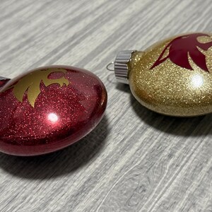 May include: Two oval-shaped holiday ornaments with metallic caps. One ornament is deep red with a gold eagle silhouette, and the other is gold with a deep red eagle silhouette. Both feature a glittery finish and a smooth, reflective surface.