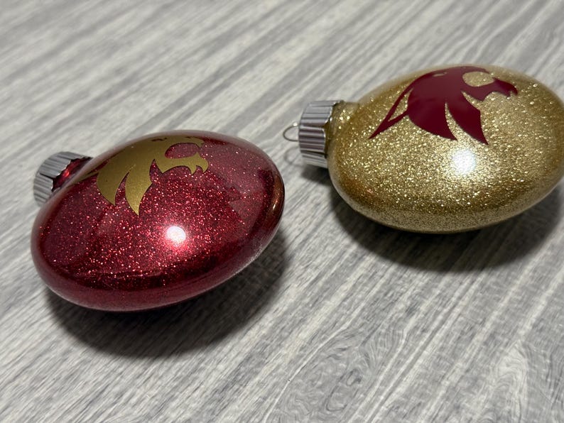 May include: Two oval-shaped holiday ornaments with metallic caps. One ornament is deep red with a gold eagle silhouette, and the other is gold with a deep red eagle silhouette. Both feature a glittery finish and a smooth, reflective surface.