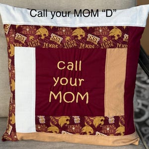 May include: A maroon and tan pillow with a white border. The pillow is decorated with a patchwork of fabric featuring the Texas State University logo and the words "Call your MOM" embroidered in gold thread.