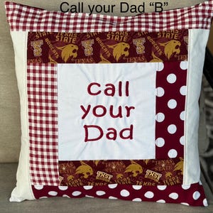 May include: A white pillow with a red and white checkered border and a red and white polka dot border. The center of the pillow has a white fabric with the words "call your Dad" embroidered in red. The pillow is decorated with a red and gold fabric with the words "Texas State Bobcats" repeated.