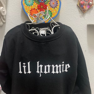 Peut inclure: Sweat-shirt noir avec un texte blanc qui dit "lil homie" dans une police gothique.