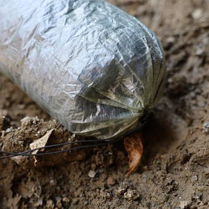 May include: A close-up of a gray, lightweight, waterproof stuff sack with a drawstring closure. The sack is lying on a dirt surface.