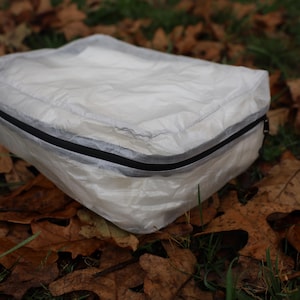 May include: A clear, white, rectangular packing cube with a black zipper. The cube is lying on a bed of brown leaves.