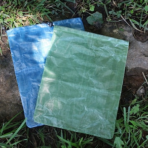May include: Two drawstring bags made of thin, translucent fabric. One bag is blue and the other is green. The bags are lying on a rock covered in grass.