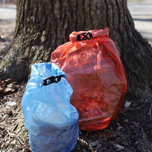 May include: Two dry bags, one red and one blue, are sitting on the ground next to a tree trunk. The bags are made of a thin, translucent material and have a drawstring closure with a clip.
