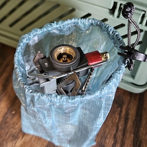 Op de afbeelding: Een verzameling kampeermateriaal, waaronder een klein fornuis en keukengerei, zit in een lichtblauwe trekkoordtas. De tas staat op een houten oppervlak, met een groene opbergcontainer op de achtergrond.