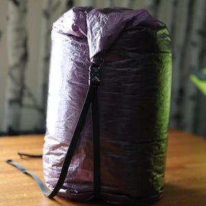 May include: A cylindrical, purple and black dry bag with a black strap and buckle closure. The bag is made of a crinkled, waterproof material and sits on a wooden surface. The bag is designed for outdoor activities.