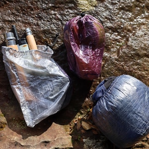 May include: Three small, lightweight, waterproof bags made of ripstop nylon. The bags are in different colors: silver, purple, and blue. The bags are lying on a rocky surface.