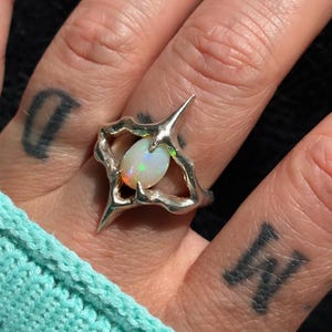 May include: A silver ring featuring an oval opal gemstone. The ring has a unique, openwork design with sharp, pointed elements. The opal displays iridescent colors. The ring is worn on a finger, with a turquoise knitted sleeve visible in the background.