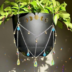 Puede incluir: Una maceta colgante negra con una calcomanía dorada de las fases lunares y cadenas plateadas adornadas con pequeñas estrellas doradas y cristales iridiscentes en forma de lágrima de color azul y transparente. Contiene una planta verde.