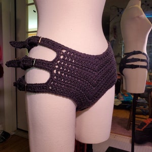 May include: A dark gray crocheted garment with a high waist and a unique design featuring three cutouts and metal rings. The garment is displayed on a white mannequin.