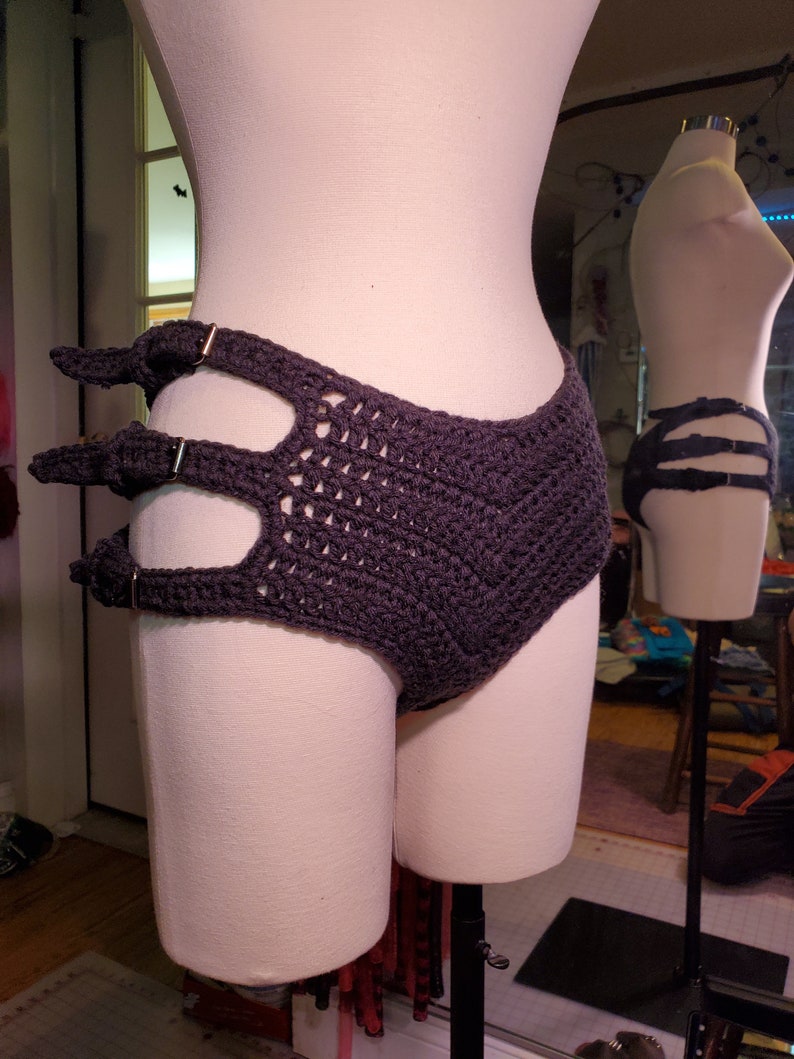 May include: A dark gray crocheted garment with a high waist and a unique design featuring three cutouts and metal rings. The garment is displayed on a white mannequin.