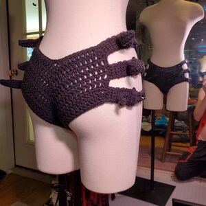 May include: A black crocheted garment with a unique design featuring multiple cutouts and pom-poms. The garment is displayed on a mannequin.