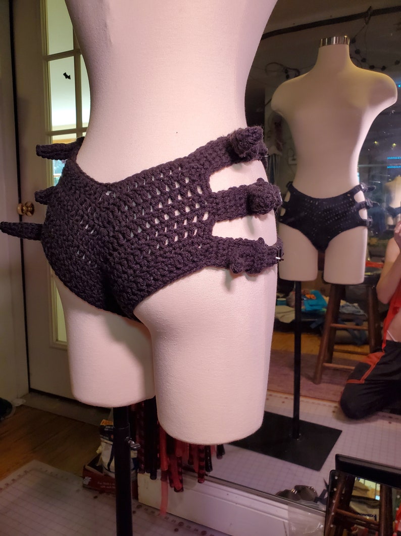 May include: A black crocheted garment with a unique design featuring multiple cutouts and pom-poms. The garment is displayed on a mannequin.