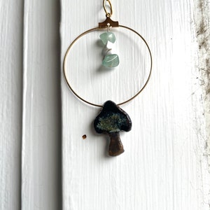 Puede incluir: Un collar de cadena dorada con un colgante circular que presenta dos piedras de color verde claro y un amuleto de hongo de cerámica con un esmalte verde oscuro y marrón.