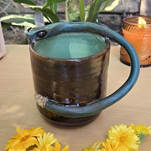 May include: A ceramic mug with a dark brown exterior and a light green interior. The handle is shaped like a green snake that wraps around the side of the mug, with the snake's head resting on the rim and its tail coiled near the base of the mug.