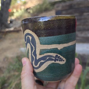 Puede incluir: Una taza cilíndrica de cerámica con un acabado esmaltado en verde oscuro y marrón. La superficie exterior muestra una ilustración de serpiente en beige claro texturizado que se enrosca alrededor del cuerpo, contrastando con el esmalte oscuro.