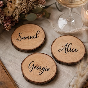 Peut inclure: Trois sous-verres circulaires en bois avec des bords en écorce naturelle, présentant chacun une surface brun clair lisse avec un grain de bois visible. Les prénoms Samuel, Alice et Georgie sont imprimés en écriture cursive noire au centre.