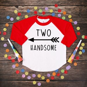 Może przedstawiać: Biała i czerwona koszulka w stylu raglan z napisem "TWO HANDSOME" i grafiką strzałki. Koszulka otoczona jest kolorowym konfetti i trąbkami na ciemnej drewnianej powierzchni. Koszulka z motywem urodzinowym.