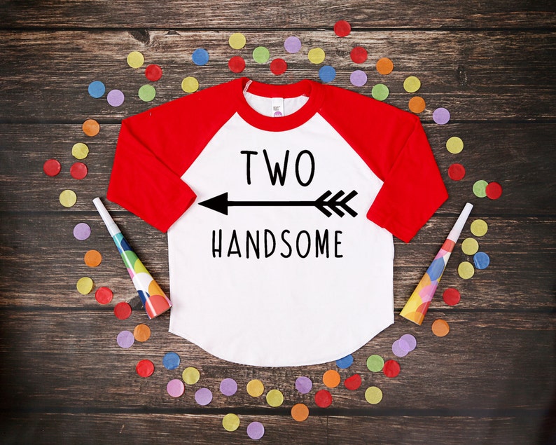Może przedstawiać: Biała i czerwona koszulka w stylu raglan z napisem "TWO HANDSOME" i grafiką strzałki. Koszulka otoczona jest kolorowym konfetti i trąbkami na ciemnej drewnianej powierzchni. Koszulka z motywem urodzinowym.
