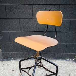 May include: A vintage industrial swivel chair with a black metal frame and a light brown wood seat and back. The chair has four wheels and a footrest.