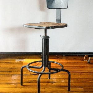 Op de afbeelding: Industriële verstelbare kruk met een zwart metalen frame en een kleine rugleuning. De zitting is van hout. De kruk heeft een cirkelvormige voetensteun en vier poten. De kruk staat op een houten vloer.
