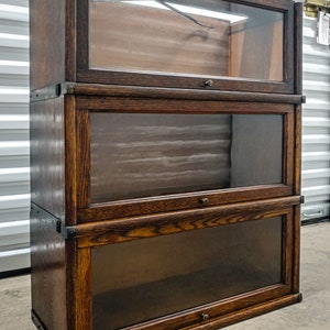 May include: A three-tiered wooden bookcase with glass doors. The bookcase is made of dark wood with a grain pattern. Each shelf has a brass knob.