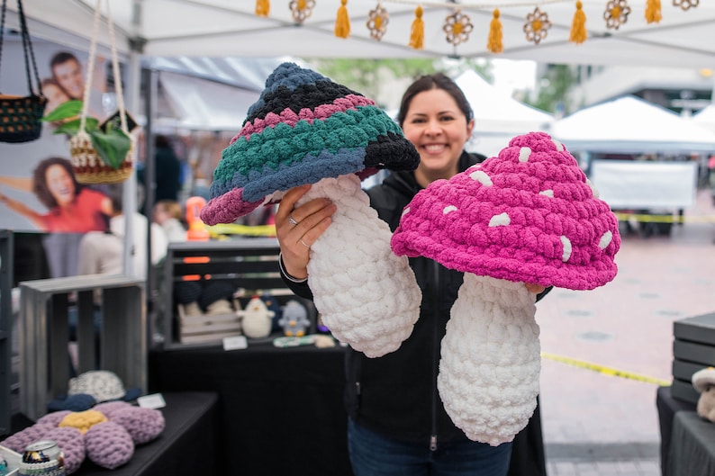 May include: Two crocheted mushroom-shaped plushies. One is a multi-colored mushroom with a white stem. The other is a pink mushroom with white spots and a white stem.