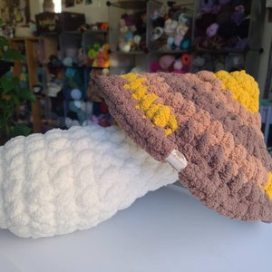 May include: A crocheted mushroom with a brown and yellow cap and a white stem. The mushroom is made with a textured yarn and has a fluffy appearance.