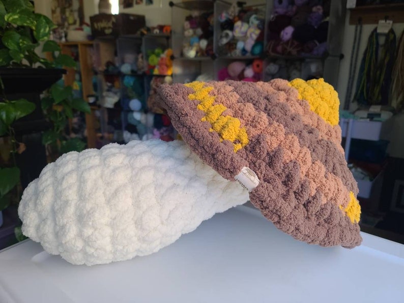 May include: A crocheted mushroom with a brown and yellow cap and a white stem. The mushroom is made with a textured yarn and has a fluffy appearance.