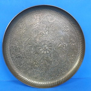 May include: A large, round metal tray with an intricate floral pattern. The tray has a raised edge and a detailed design of flowers and leaves. The metal has a slightly tarnished appearance, giving it a vintage look. The tray is likely used for serving or display.