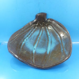 May include: A handmade ceramic dish in the shape of a fig. The dish features a dark brown exterior with a turquoise interior and vertical grooves. The dish is on a blue background.