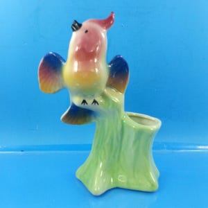 Może przedstawiać: Ceramiczna figurka ptaka o kolorowym designie. Ptak ma różową głowę, żółtą pierś i niebieskie skrzydła. Siedzi na zielonym pniu drzewa z małym otworem, prawdopodobnie doniczce lub wazonie. Na niebieskim tle.