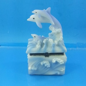 May include: A white and blue decorative box featuring three dolphins leaping from ocean waves. The box has a hinged lid and a black clasp. The dolphins are white with black eyes, and the waves are detailed with a foamy texture.