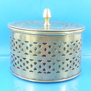 May include: A round, silver-toned metal container with a lid. The container has a decorative pierced design around the sides. The lid has a floral pattern and a gold-colored knob. The container sits against a blue background.