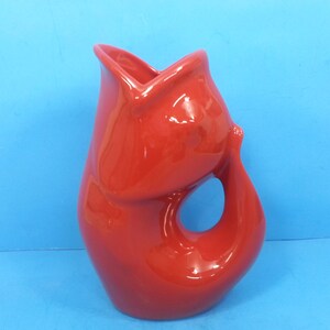 May include: A glossy, red ceramic jug with a unique, organic shape. The jug features a handle with a large opening and a flared top. The bright red colour and unusual design make it a striking decorative piece.