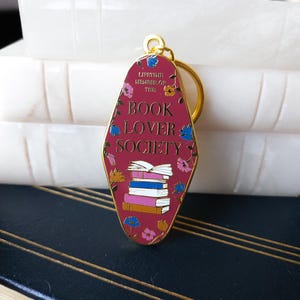May include: A red metal keychain shaped like a motel sign with the text "Lifetime Member of the Book Lover Society" and a stack of books with floral accents.