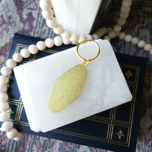 May include: A gold motel key chain with a sparkly finish. The key chain is on top of a white book with a blue cover.