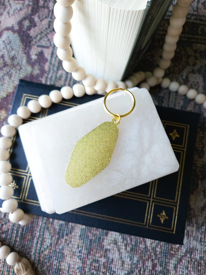 May include: A gold motel key chain with a sparkly finish. The key chain is on top of a white book with a blue cover.