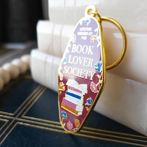 May include: Gold-toned keychain with a pink and red enamel charm shaped like a motel sign. The charm features a stack of books, flowers, and the text "Lifetime Member of the Book Lover Society".