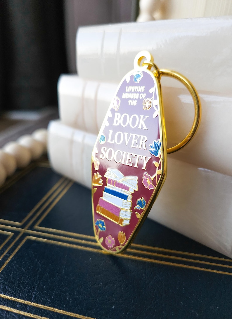 May include: Gold-toned keychain with a pink and red enamel charm shaped like a motel sign. The charm features a stack of books, flowers, and the text "Lifetime Member of the Book Lover Society".