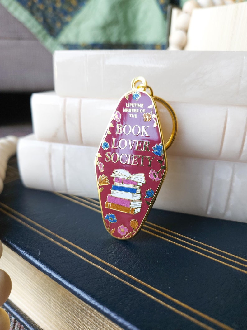 May include: A gold-colored keychain with a red motel-style sign that says "Lifetime Member of the Book Lover Society". The sign is decorated with a stack of books and floral designs.