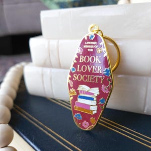 May include: A red metal keychain shaped like a motel sign with the text "Lifetime Member of the Book Lover Society" and a stack of books in the center. The keychain has gold accents and floral details.