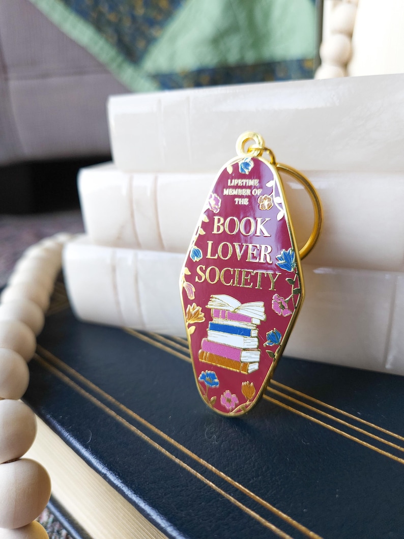 May include: A red metal keychain shaped like a motel sign with the text "Lifetime Member of the Book Lover Society" and a stack of books in the center. The keychain has gold accents and floral details.